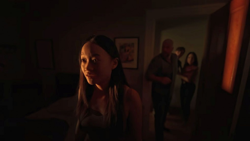 A girl stands in a dark room looking scared, her family in the doorway.