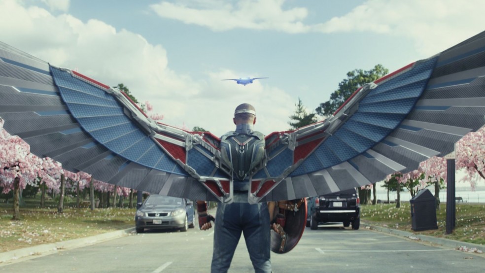 A superhero with mechanical wings stands in a road flanked by pink blossom.