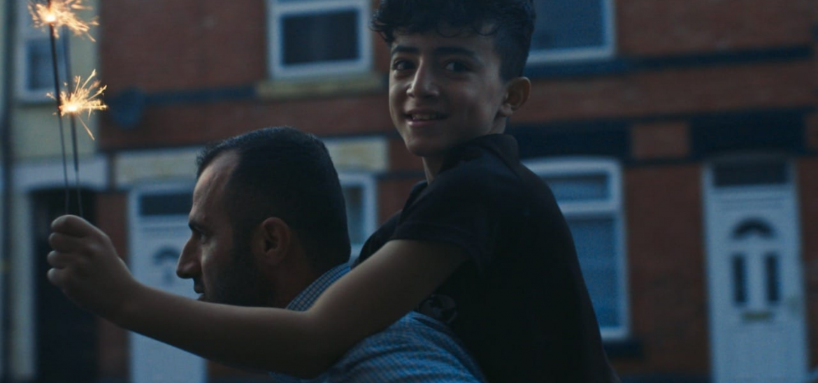 A man carried a boy on his back, who holds two lit sparklers and smiles.