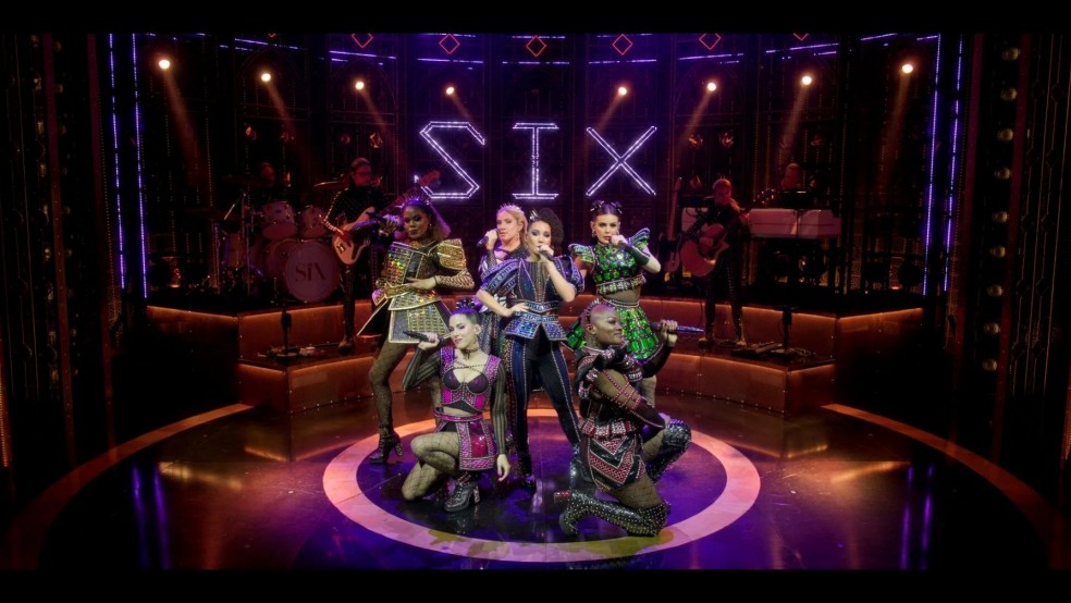 Six women wearing Tudor inspired clothes pose with microphones on a stage.