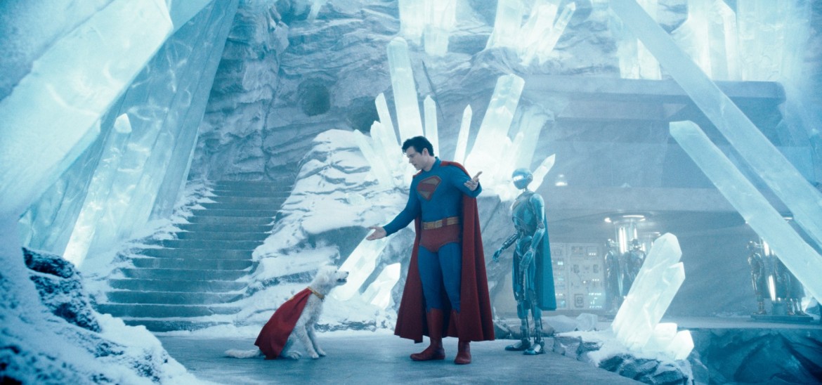 A man in a superhero costume talks to a dog wearing a cape in an icy cave.