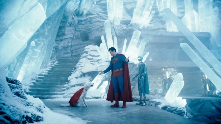 A man in a superhero costume talks to a dog wearing a cape in an icy cave.