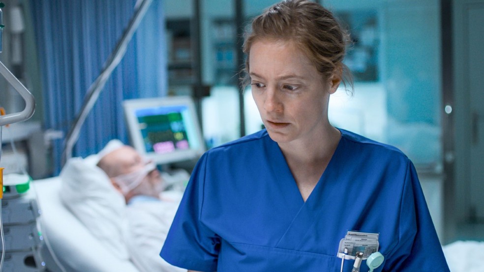 A woman wearing a nurse uniform stands with her back to a hospital bed.