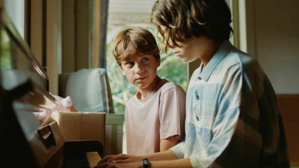 Two adolescent boys sit at a piano, one watching the other play.