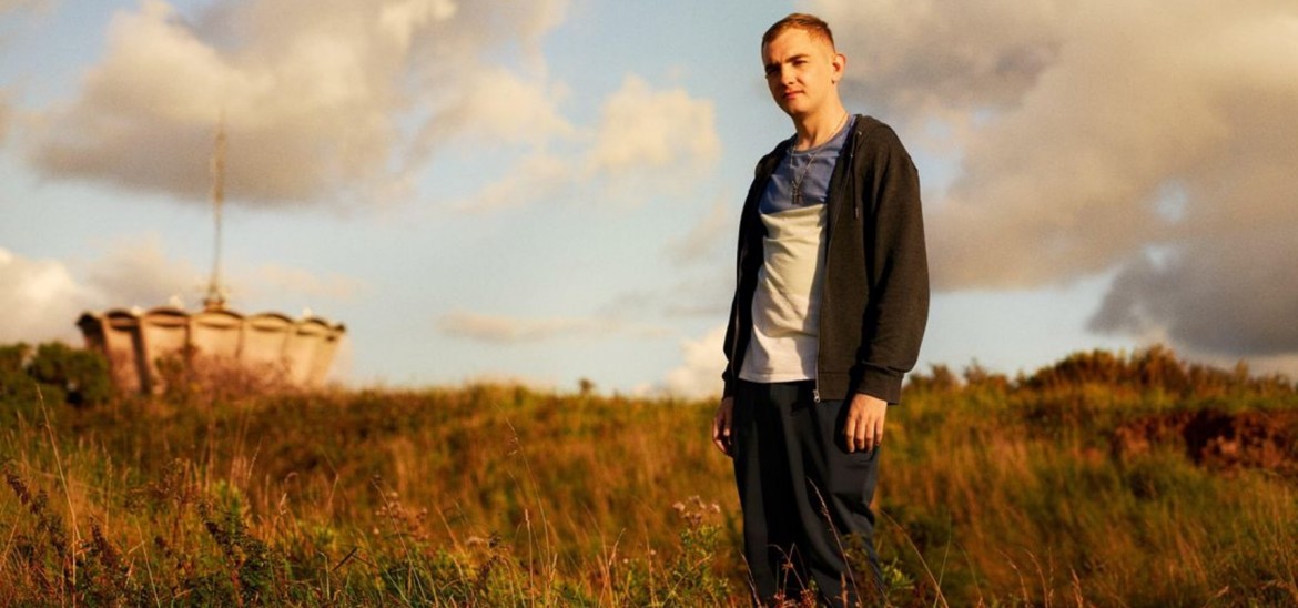 A teenage boy with short blond hair and a zip up hoodie stands in a field.