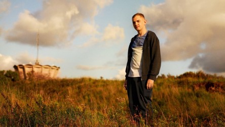 A teenage boy with short blond hair and a zip up hoodie stands in a field.