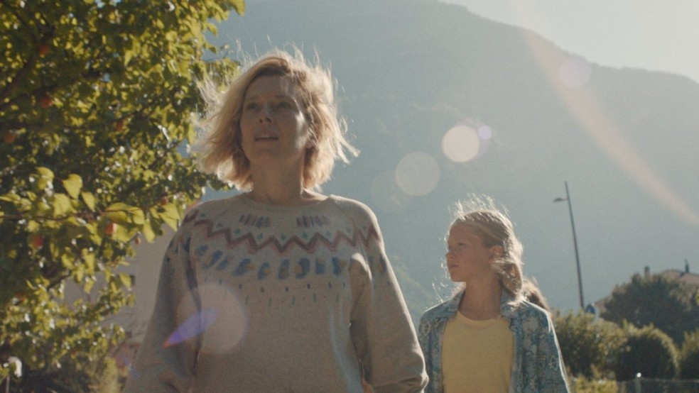 A woman and a young girl walk along a sunny road in front of a mountain.