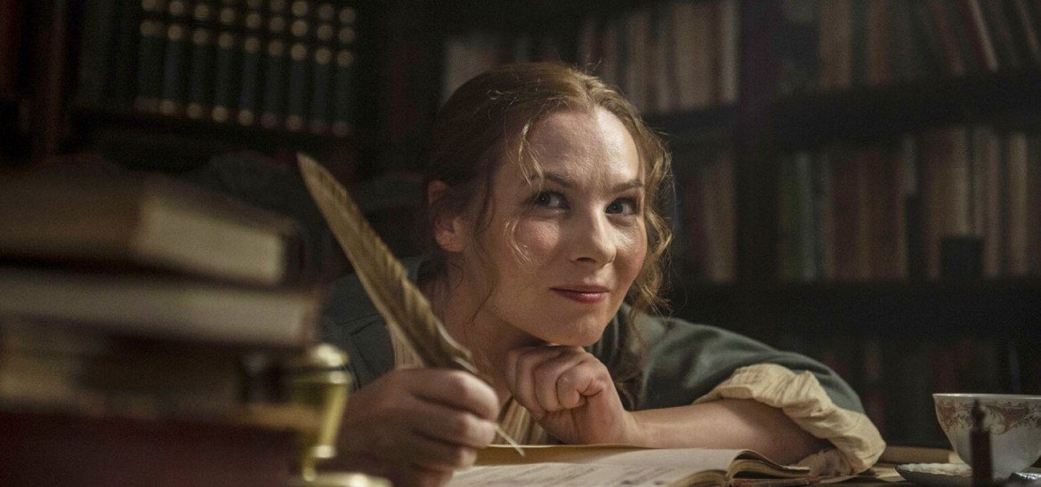 A woman writes in a book in a library with a feather quill, smiling.
