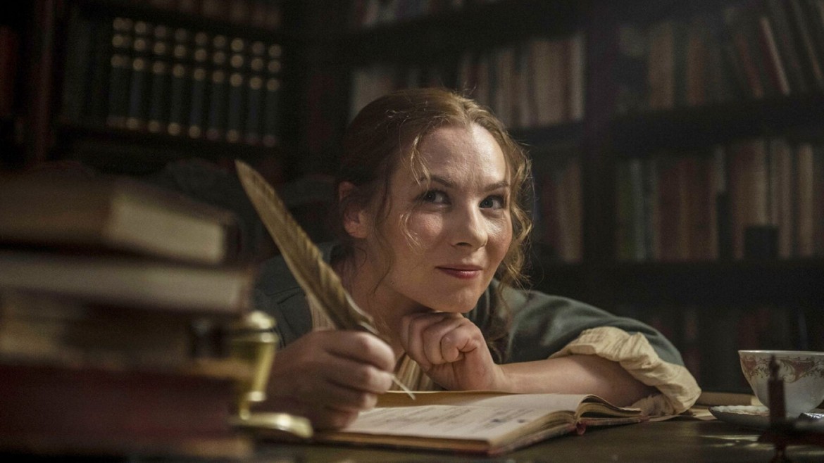 A woman writes in a book in a library with a feather quill, smiling.