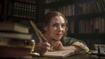 A woman writes in a book in a library with a feather quill, smiling.