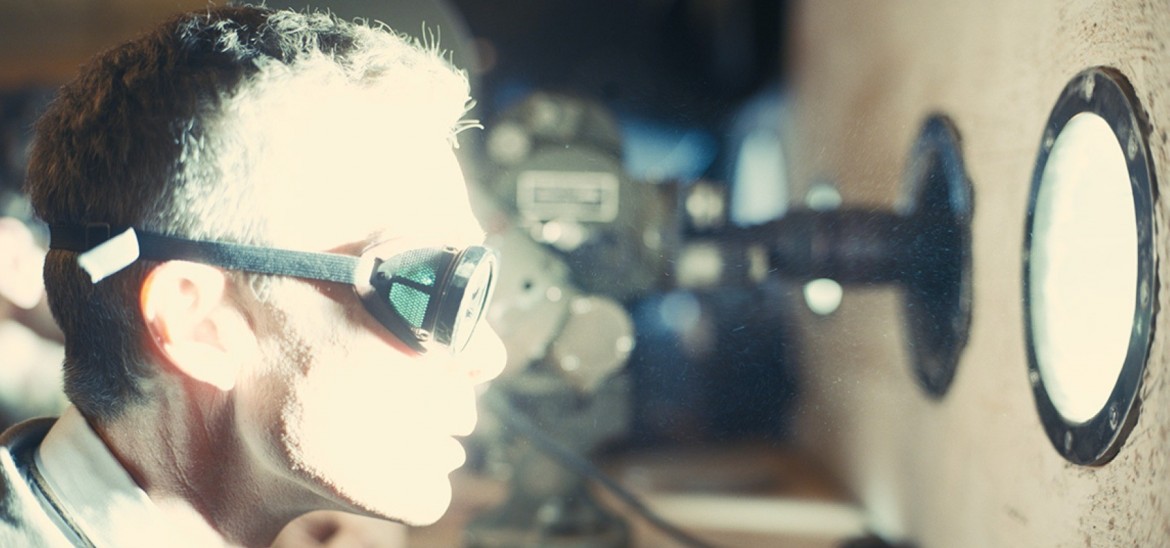 A man wearing goggles looks through a small bright circular window.