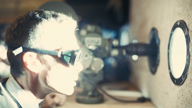 A man wearing goggles looks through a small bright circular window.