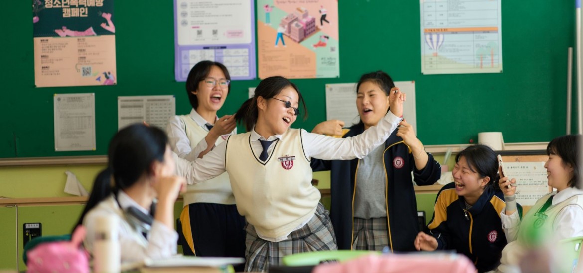 A girl dances in a classroom surrounded by a group of other girls laughing.