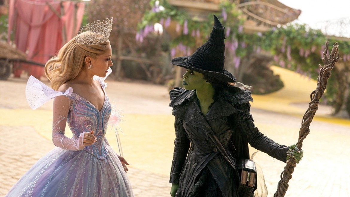 A blonde woman in a pink dress faces a green woman in a black witch outfit.