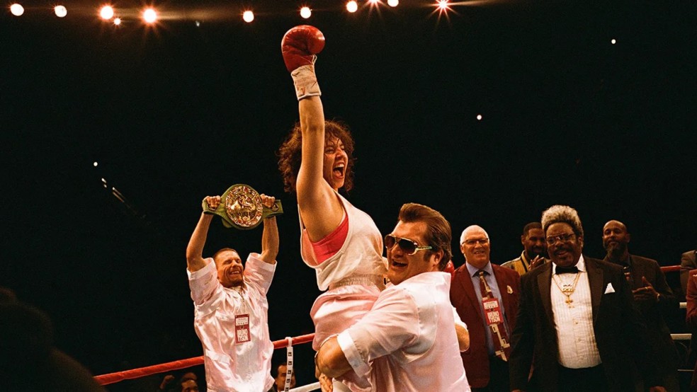A female boxer is triumphantly lifted into the air in a boxing ring.