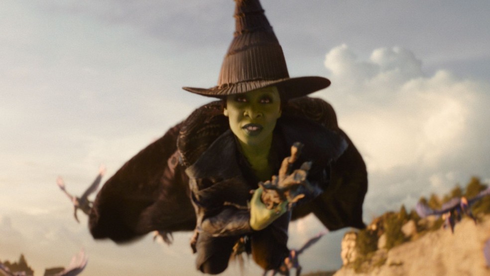 A green-skinned witch with black clothing flies on a broomstick.