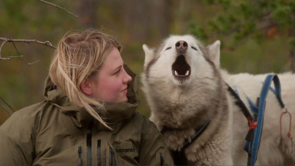 A young woman in a heavy coat sits next to a husky who is howling