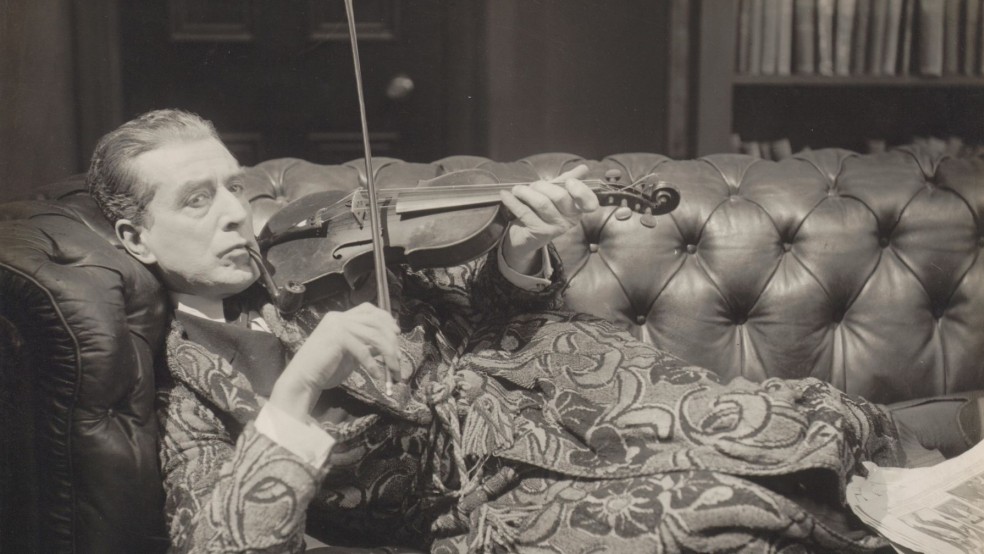 A black and white image of a man reclining on a sofa playing the violin.