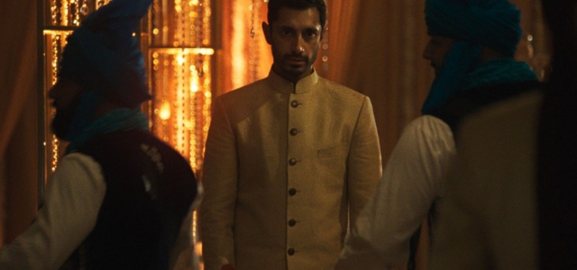 A man in a sherwani stares ahead as people move past him in a dark room.