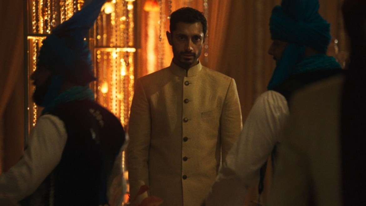 A man in a sherwani stares ahead as people move past him in a dark room.