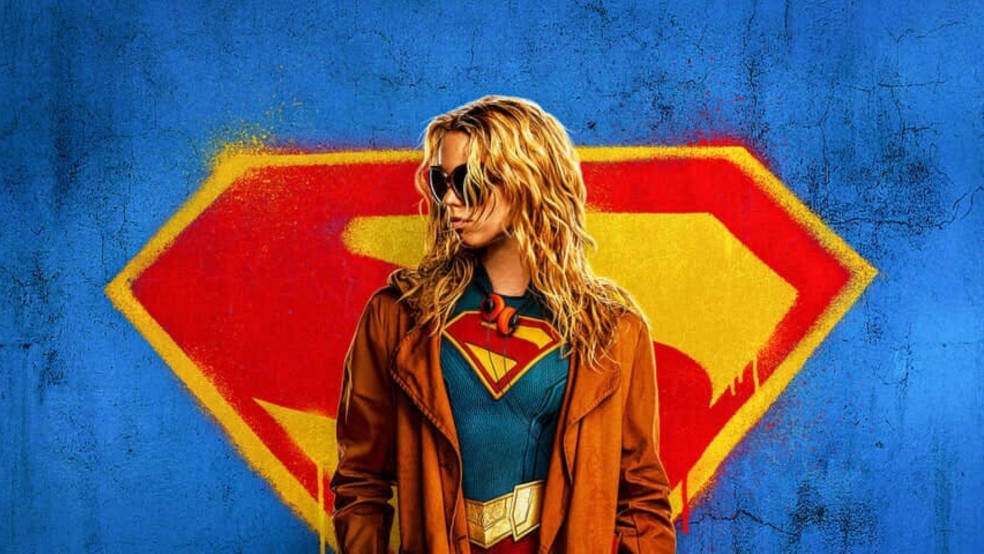 A girl in a superhero outfit stands in front of a giant spray-painted S
