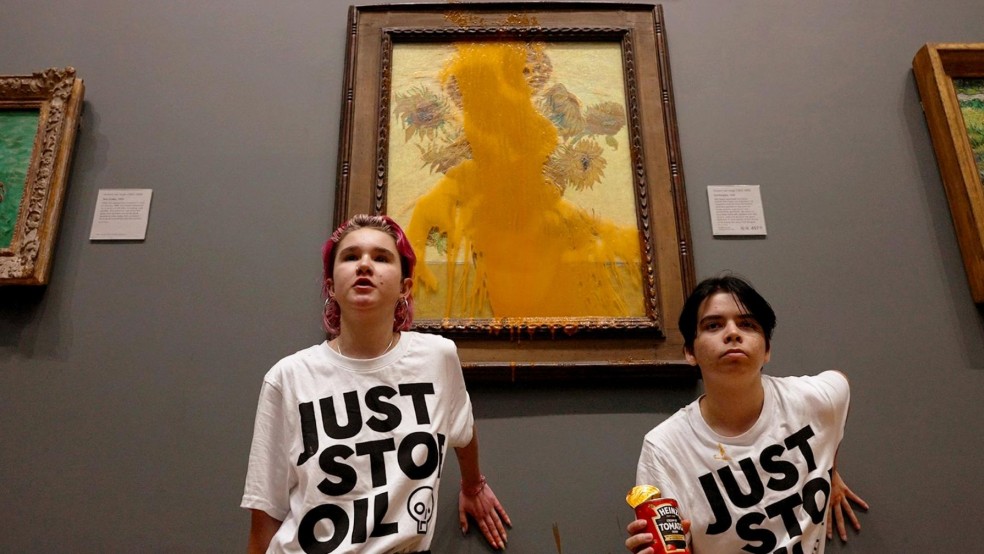 Two young people stand in front of a painting with soup splashed over it.