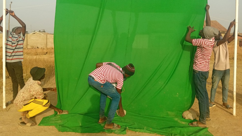 A group of young people hang a green screen from a frame outdoors.