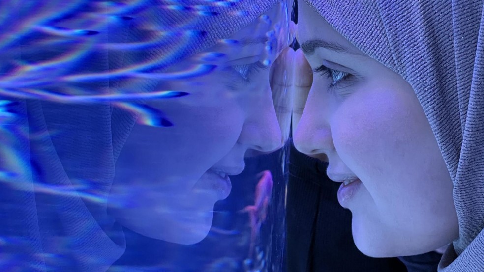 A woman wearing a headscarf leans her head against glass in an aquarium.