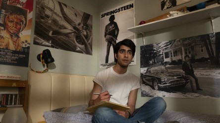 A teenage boy sits on a bed writing, surrounded by Springsteen posters.
