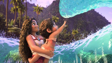 A woman holds a child in her arms on a beach, as a wave touches her hand.